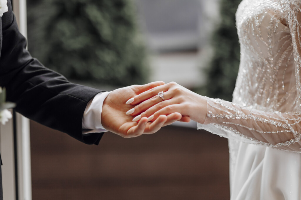 The Man Gently Holds The Bride's Hand With A Beautiful Golden Wedding Ring. Enlarged Image Of Newlyweds' Hands. Wedding Concept. The Groom Gently Touches The Bride