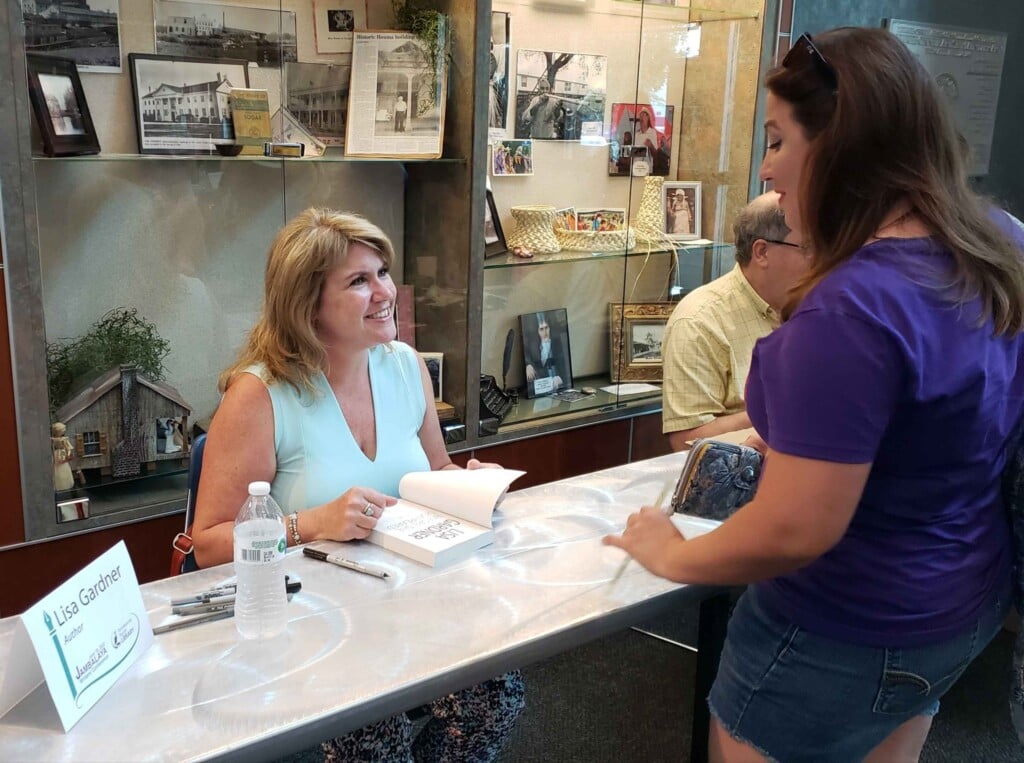 Jambalaya Writer's Conference 2022 - Woman sitting at a table and smiling while opening book she is about to sign for woman at the conference