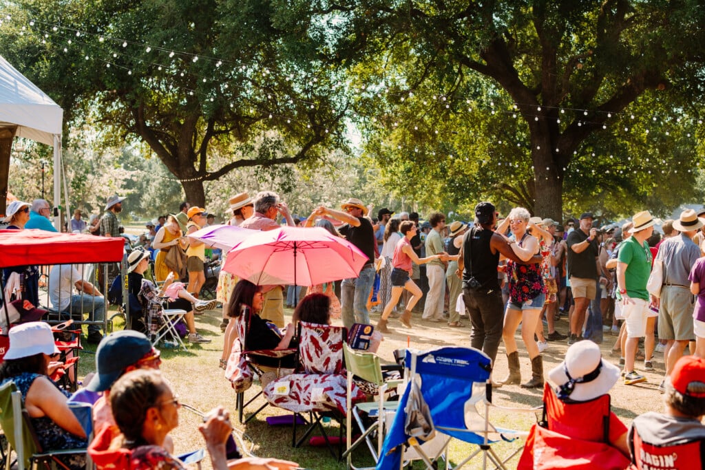 2024 Festivals Acadiens et Créoles showing people with chairs, umbrellas and tents as they listen and dance to music. Trees in the background with strings of lights hanging from the trees