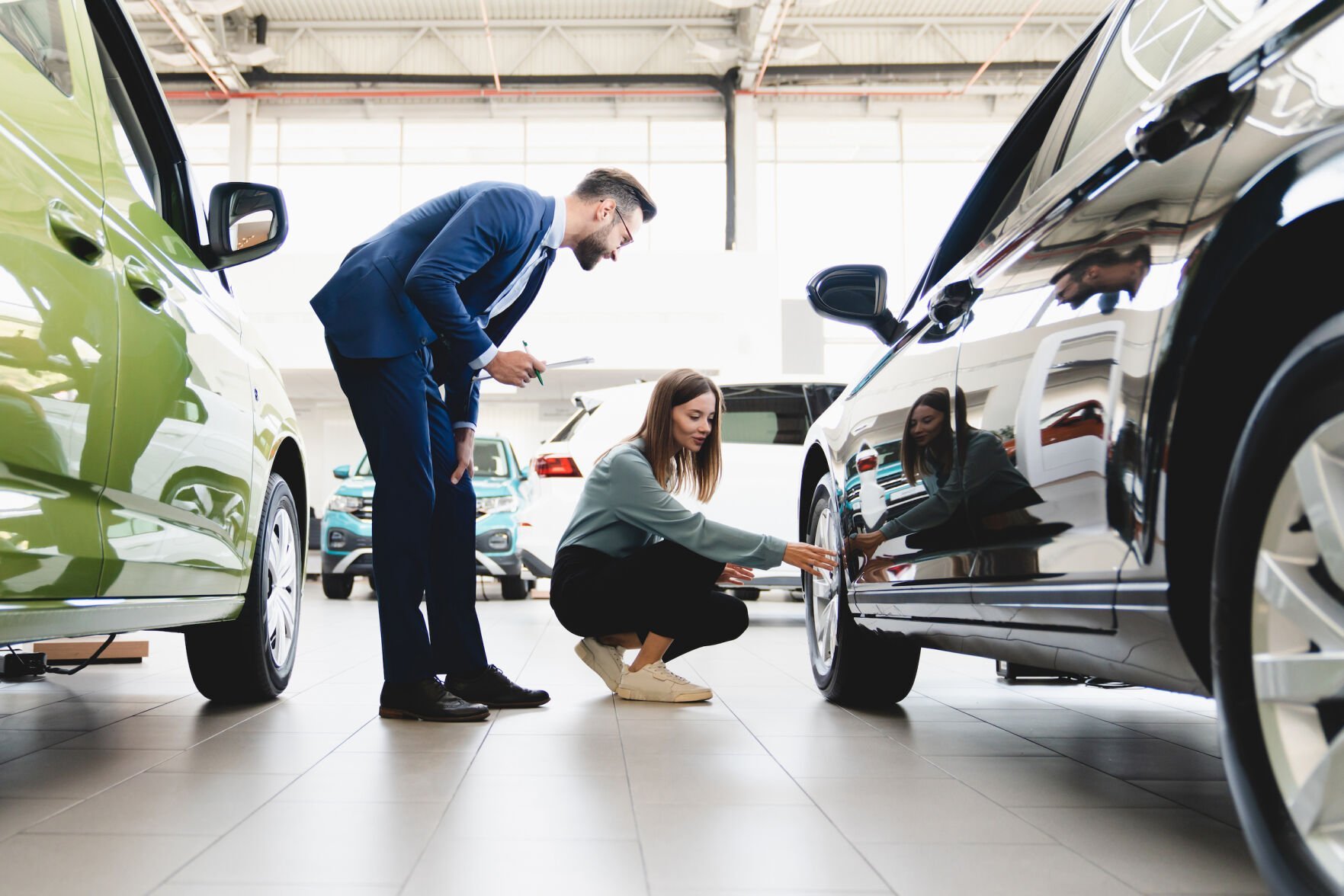 <p>Beautiful young caucasian female client customer choosing new car, trying checking its options, tire, wheels while male shop assistant helping her to choose it at dealer auto shop</p>