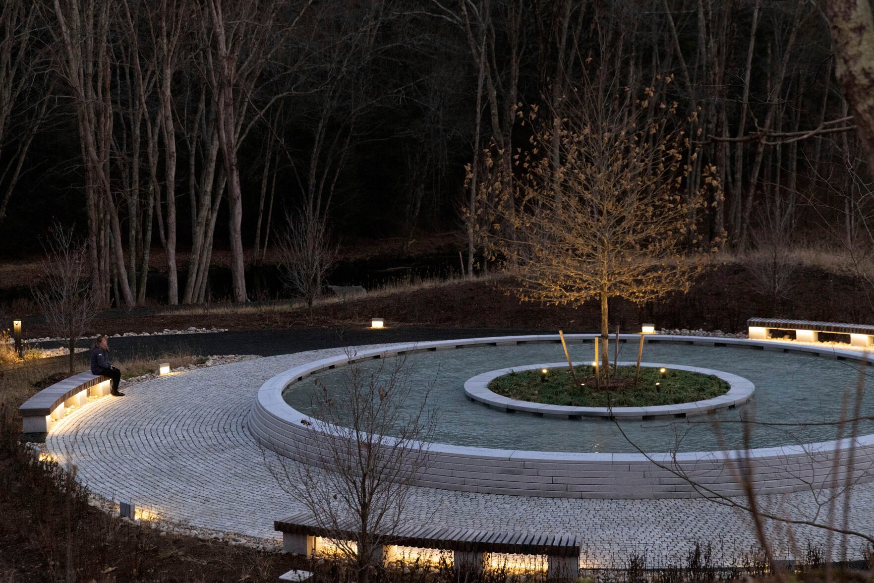 <p>A visitor to the Sandy Hook Permanent Memorial sits on one of the benches during twilight Dec. 5 in Newtown, Conn. The names of the 20 children and six educators killed a short distance away at Sandy Hook Elementary School 10 years ago are engraved in concrete around a memorial pool with a sycamore tree in the middle.</p>