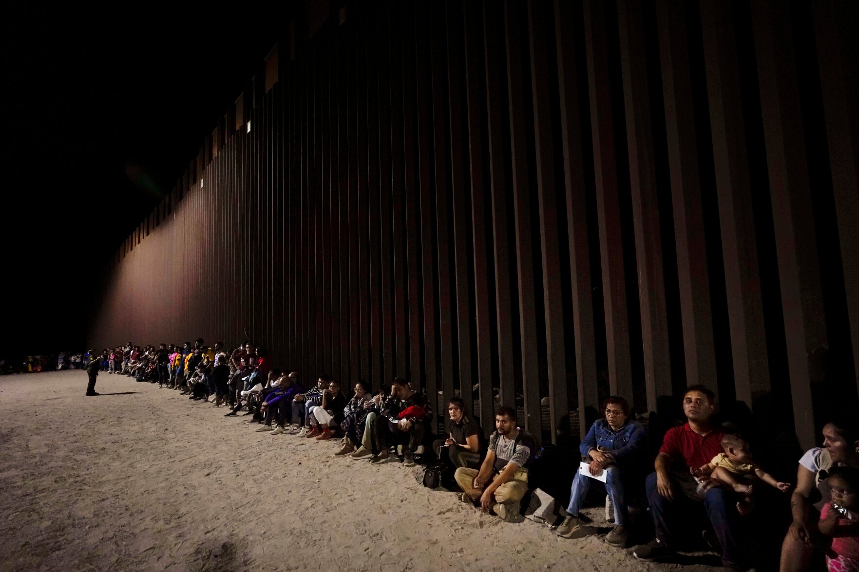 <p>Migrants wait along a border wall Aug. 23 after crossing from Mexico near Yuma, Ariz. President Joe Biden's administration announced in early January that it would admit up to 30,000 people a month from Cuba, Haiti, Nicaragua and Venezuela for two years with authorization to work when they apply online.</p>