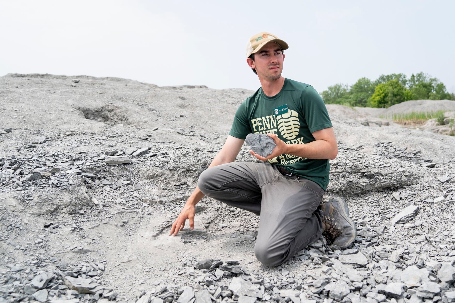 <p>Jonathan Hoag, site manager at Penn Dixie Fossil Park and Nature Reserve, kneels for a portrait with a fossil he discovered, gesturing to the rock where he found the fossil at the Penn Dixie site in Hamburg. </p>