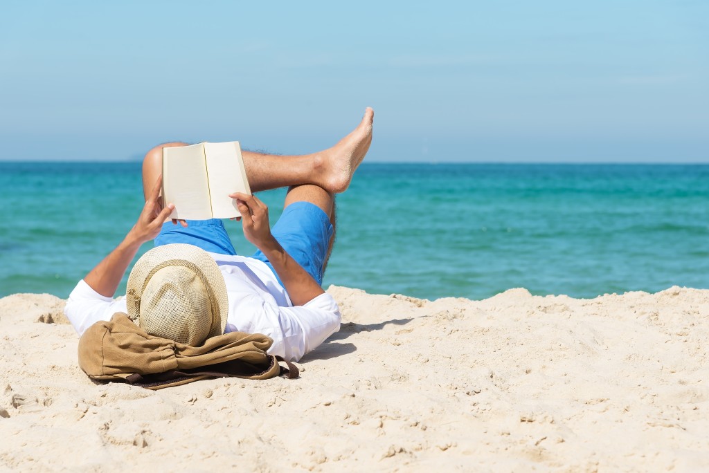 A Handsome Man Relaxing On A Book Reading On The Beach.