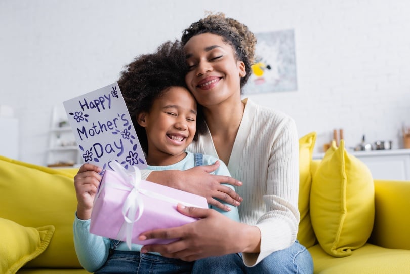 Pleased African American Woman Holding Gift Box And Embracing Daughter With Happy Mothers Day Card