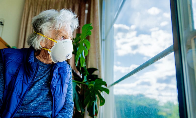 Wide Angle Low Shot Distraught Elderly Senior Caucasian Woman Looking Out The Window Feeling Loneliness Wearing An N95 Protective Face Mask To Prevent The Spread Of Covid Sars Ncov 19 Coronavirus Swine Flu H7n9 Influenza Illness During Cold And Flu Season