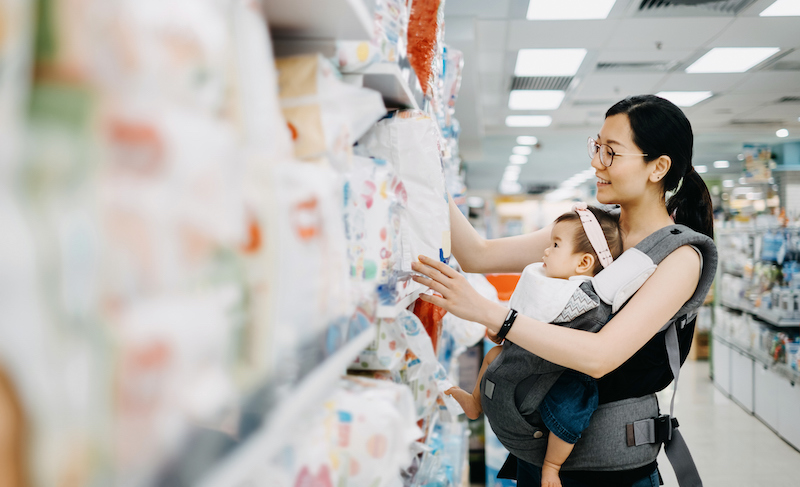 Young Asian Mother Carrying Cute Baby Girl Shopping For Baby Product In A Shopping Mall And Is Looking At A Variety Of Diapers