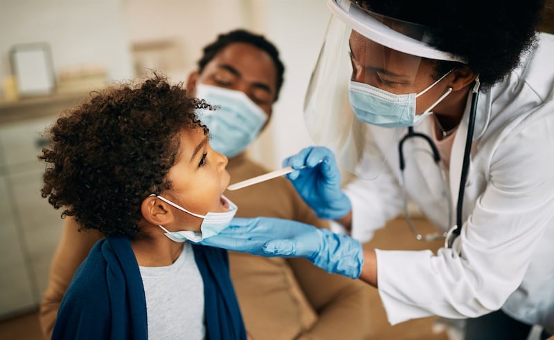 African American Doctor With Face Mask Examining Boy's Throat During A Home Visit.