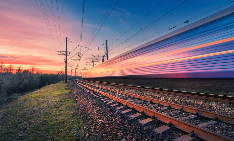 High Speed Passenger Train In Motion On Railroad At Sunset. Blurred Modern Commuter Train. Railway Station And Colorful Sky. Railroad Travel, Railway Tourism. Industrial Landscape. Transportation