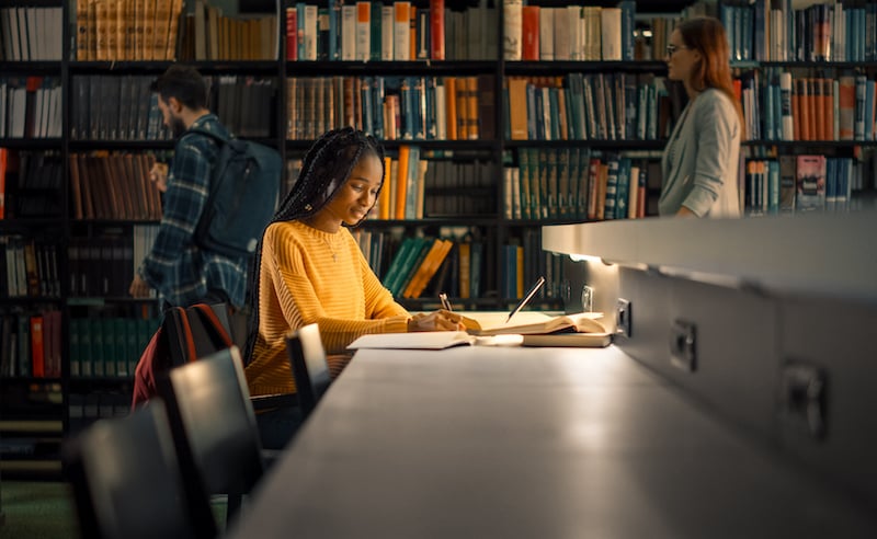 University Library: Gifted Black Girl Uses Laptop, Writes Notes For The Paper, Essay, Study For Class Assignment. Students Learning, Studying For Exams College. Side View Portrait With Bookshelves