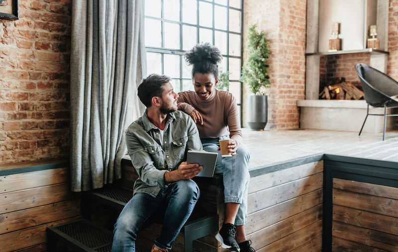 Multiracial Couple In Modern Loft Using Technologies. Couple Using Digital Tablet For Smart Home Apps, Electronic Banking And Playing Video Games Together.