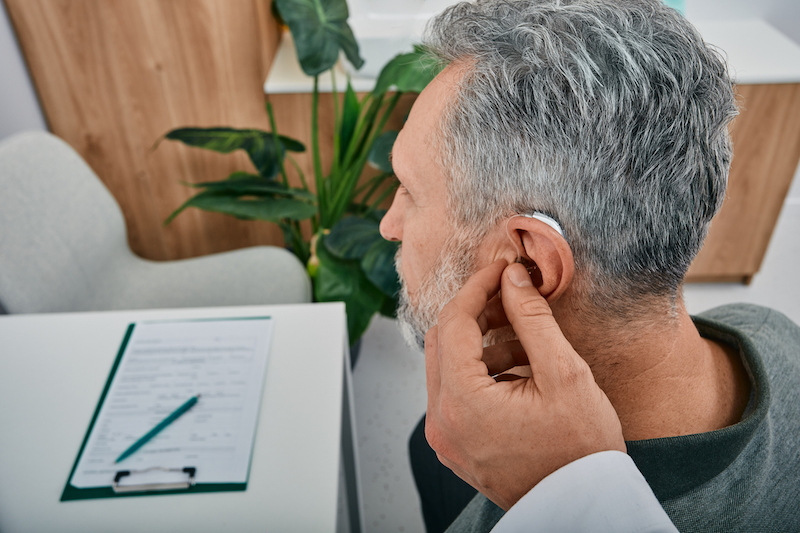 Audiologist Fits A Hearing Aid On Deafness Mature Man Ear While Visit A Hearing Clinic. Hearing Solutions For Older