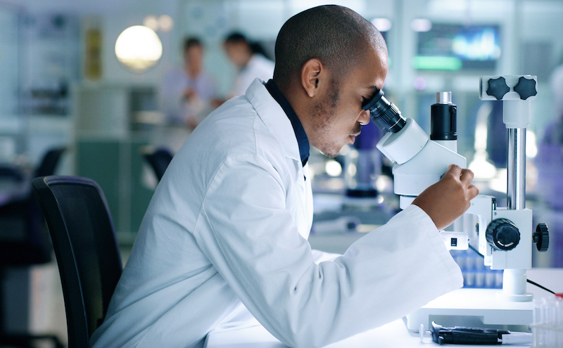 Research Scientist Analyzing A Sample, Looking Into A Microscope, Conducting An Experiment. Male Biologist Or Chemist Working On A Futuristic Medical Development In A Laboratory.