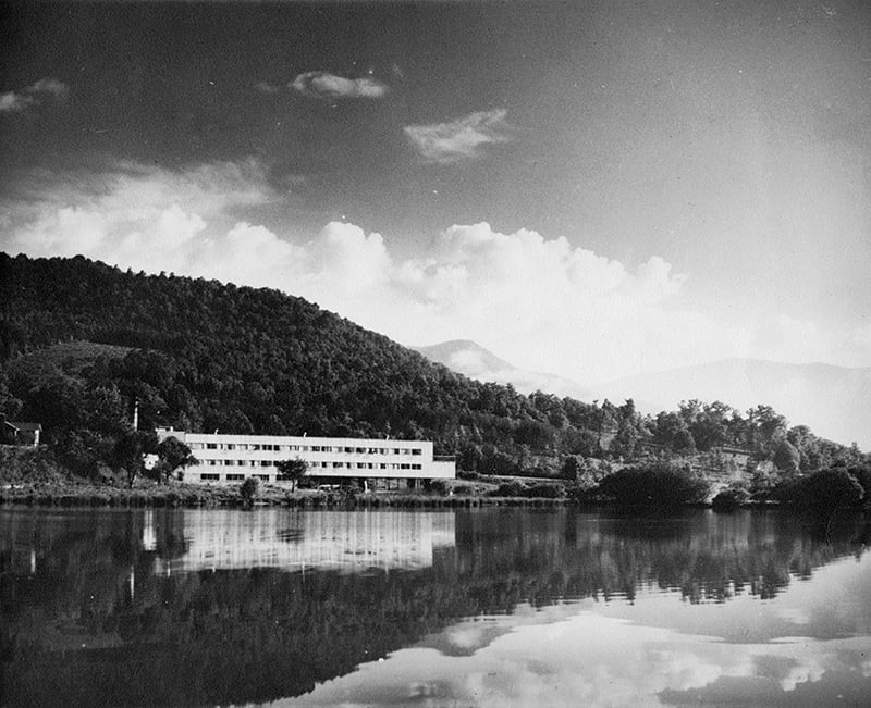 Black and white photograph of the Black Mountain College studies building overlooking Lake Eden. Cloudy sky with dark mountain curving down from left to right with light colored modern building on edge of lake and reflected back in the lake surface.