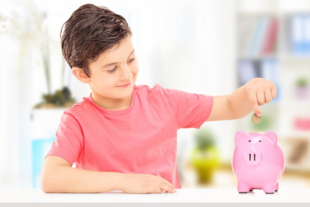 Boy,inserting,coins,into,a,piggybank,seated,at,table,indoors