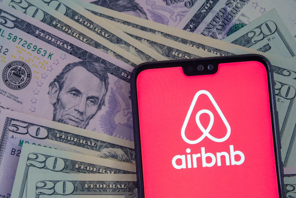 Optimize your Airbnb listing concept: Airbnb App Logo Seen On The Screen Of Smartphone, Placed On Dollar Bills.