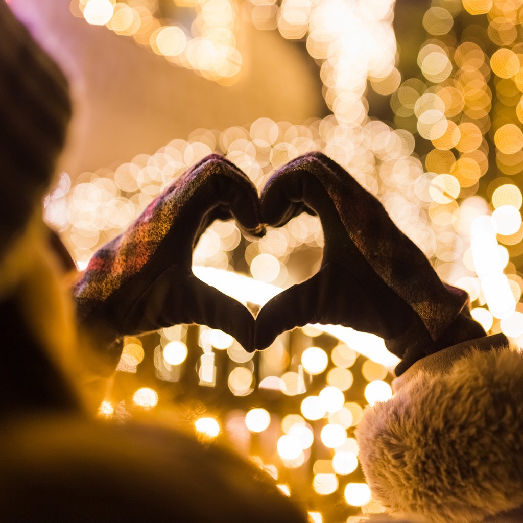 Rear View Of Woman Making Heart Shape With Her Hands Against House Decorated With Christmas Lights.