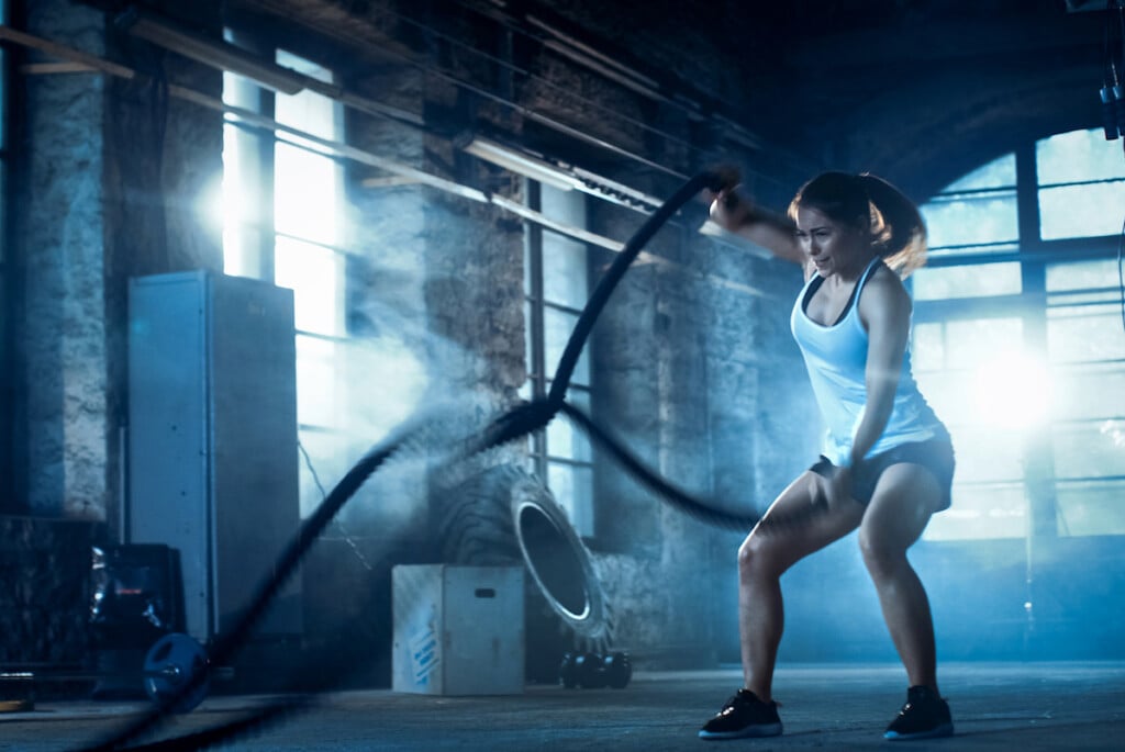 Athletic Female In A Gym Exercises With Battle Ropes During Her Workout/ High Intensity Interval Training. She's Muscular And Sweaty, Gym Is In Deserted Factory. Cold Ambient.