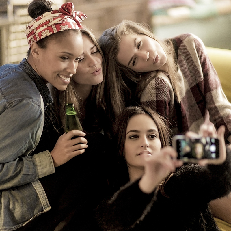 Four Young Women Sitting On A Sofa, Smiling, Taking A Selfie, Holding Beer Bottles.