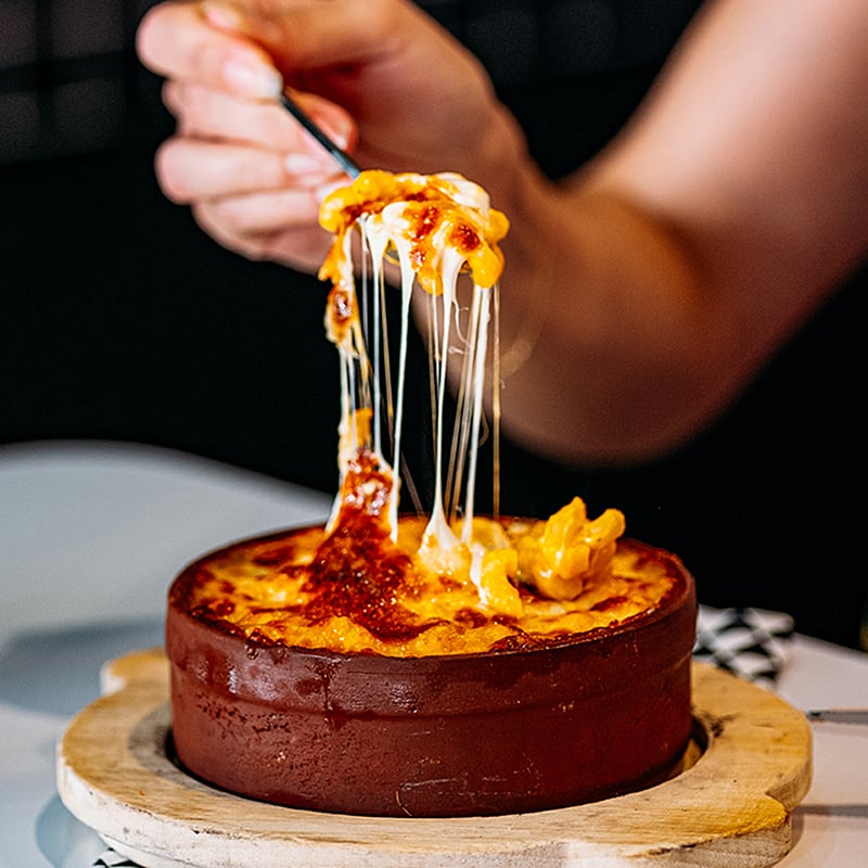 A hand holding a fork full of mac & cheese with strings from the cheese stretched from the bowl it was pulled from.