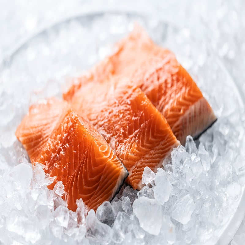 Three Slices Of Fresh Salmon, Placed On A White Plate Filled With Crushed Ice. The Salmon's Bright Orange Color Contrasts With The Cold, Icy Background, And The Whole Scene Is Completed With A Small Sprig Of Rosemary, Suggesting Culinary Preparation.