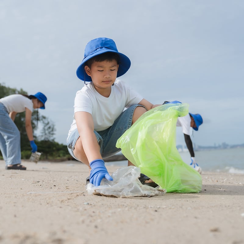 Boy Participates In Beach Cleanup Activity On A Sunny Day With Volunteers Collecting Litter And Promoting Environmental Awareness