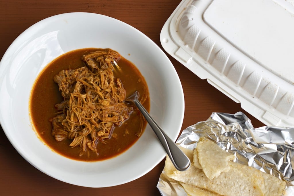 a bowl of shredded chicken surrounded by a sea of deeply red-brown mole sauce with a side of soft tortillas
