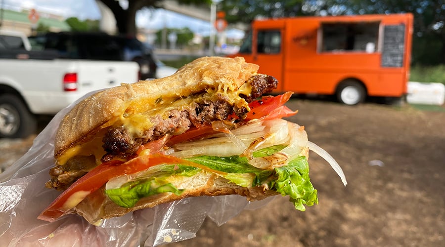 big bite taken out of a tomato- and lettuce-topped smash burger in front of orange food truck