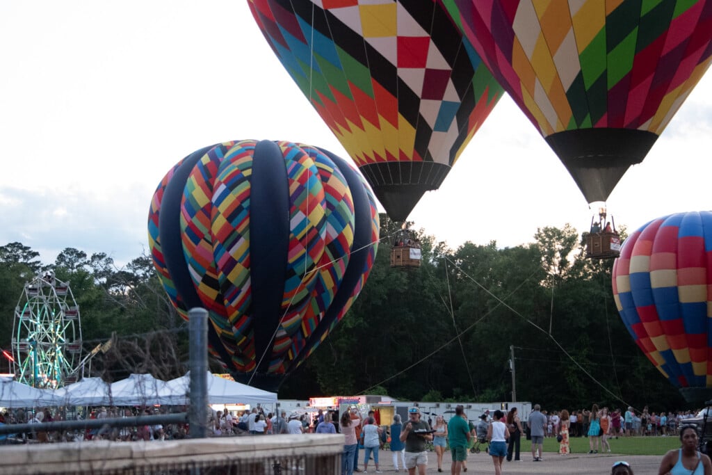 Image from the 2025 Washington Parish Balloon Festival showing four air balloons in frame, two on the ground and the two in the middle in the air right at the tree line. The air balloons surround the festival grounds with people walking, tents and a ferris wheel