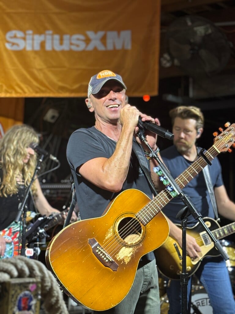 Kenny Chesney at Flora-Bama No Shoes Radio Concert on stage holding microphone with guitar strapped around him