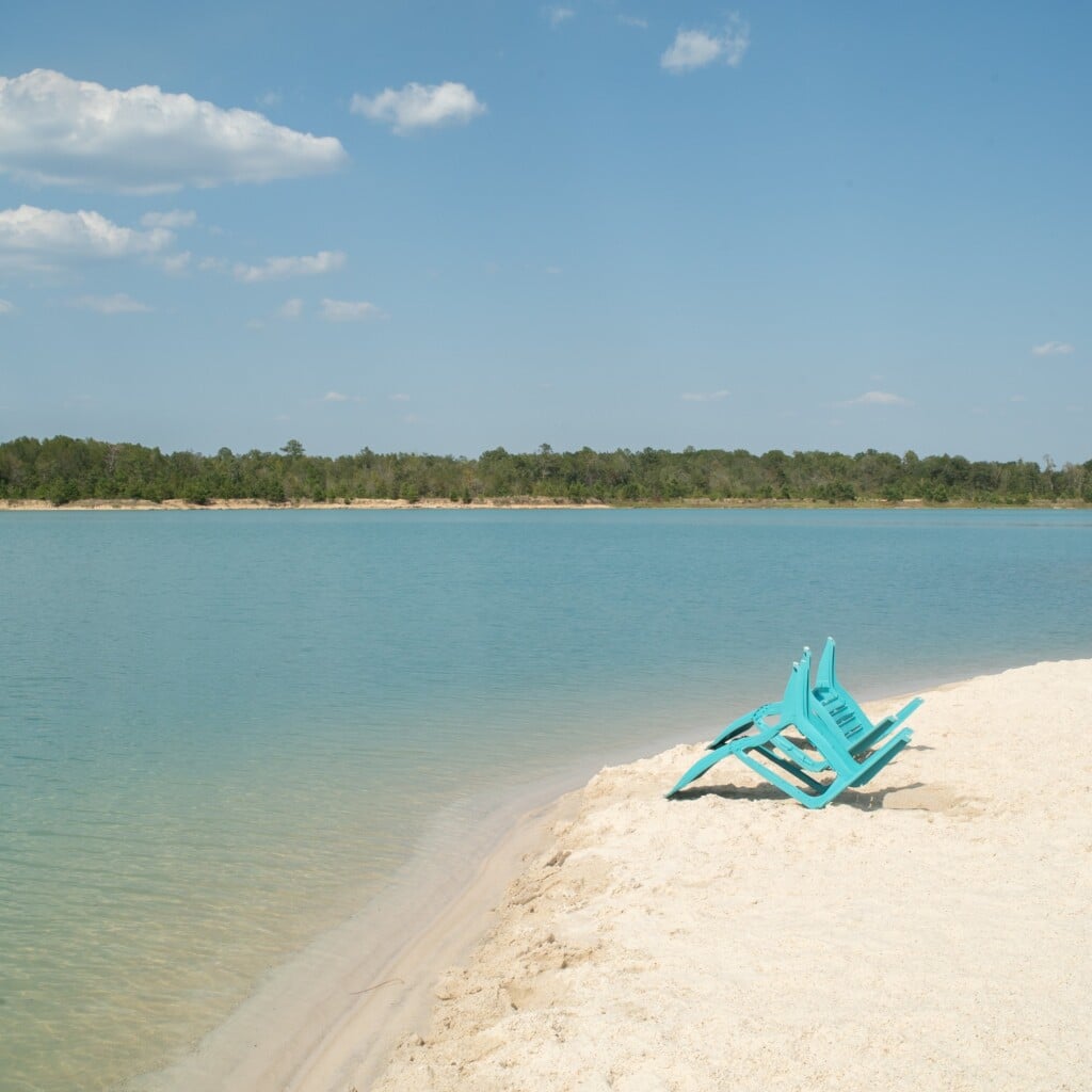 Lake Isabel at Lake Isabel Farm. White sand beach in the right corner with two blue chairs surrounded by blue open lake water with tree line in the back and blue sky with some clouds