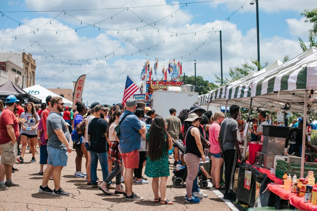 Louisiana Peach Festival: Crowd of people standing in line at tent with flags and tents in the background along the street