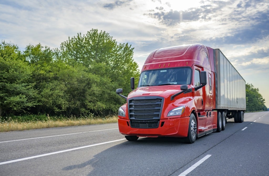 Red Big Rig Long Haul Semi Truck With Black Grille Transporting Cargo In Dry Van Semi Trailer Running On The Wide Highway Road