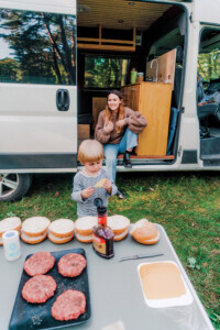 Woman Cooking Burger On Grill In Forest With Little Boy