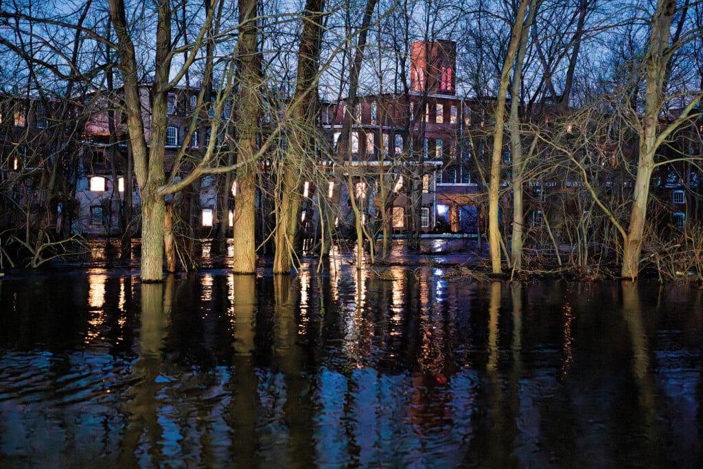 A mill building is pictured at sunset, with trees and a flooded river in the foreground.