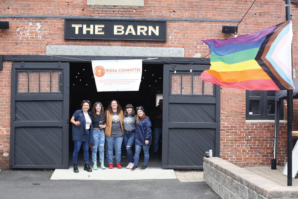 Five women stand in the doorway of a brick building with a rainbow flag in the foreground.