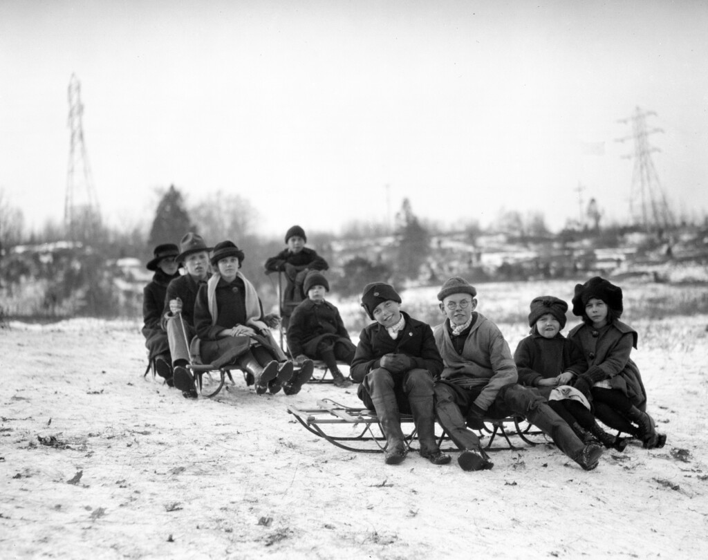 A black-and-white photo of children bundled up for winter sitting on sleds.