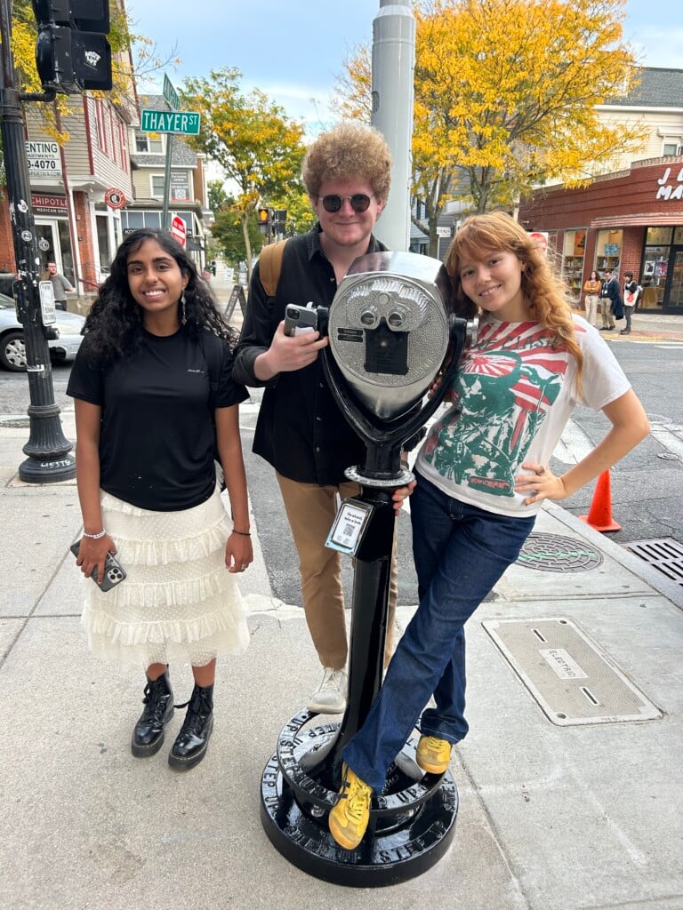 Three students pose with a binocular-style tower viewer on the sidewalk of a city intersection.