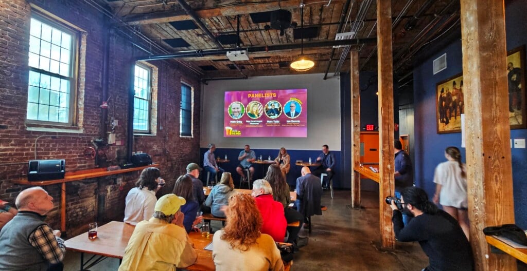 A crowd watches a panel of speakers in an industrial, brick-walled space.