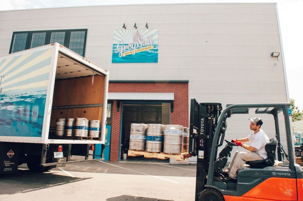 The loading dock outside a brewery, with a man driving a forklift and kegs in the foreground.
