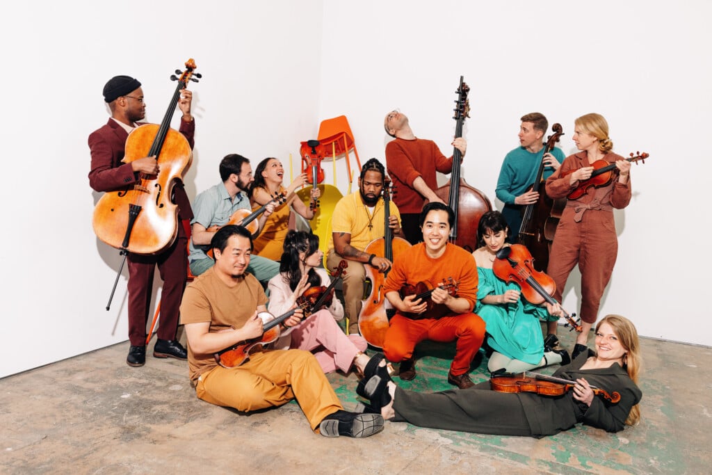 A musical ensemble featuring about a dozen musicians holding instruments and wearing bright colors.