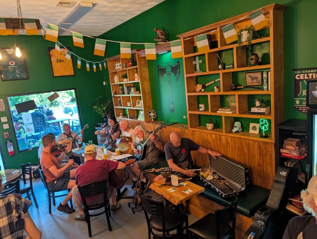Musicians sit around a table in a taproom with green walls and Irish flag decor.