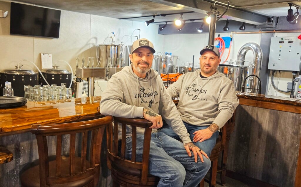 Two men in baseball caps and Uncommon Pair Brewing hoodies sit at a bar.