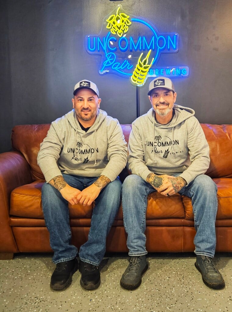 Two men sit on a couch in front of a neon sign that reads "Uncommon Pair Brewing."
