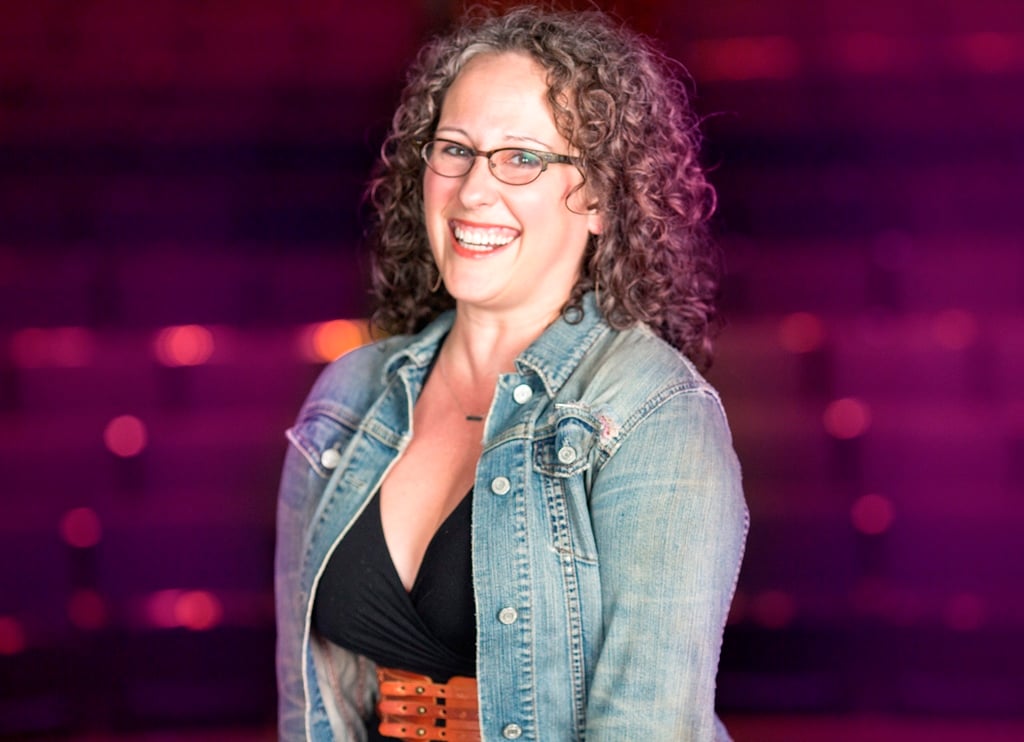A woman with curly hair and glasses wearing a jean jacket.