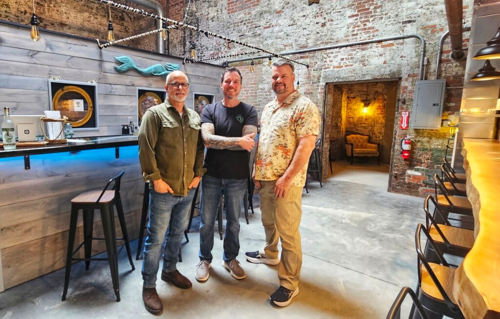 Three men stand in front of a bar with red brick walls and an industrial feel.