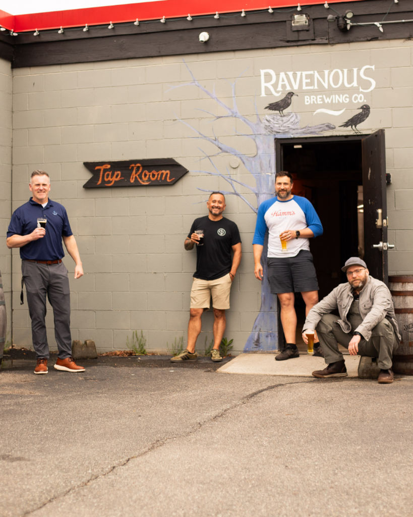 Three men holding pints of beer standing and one sitting outside the Ravenous Brewing Co. taproom.