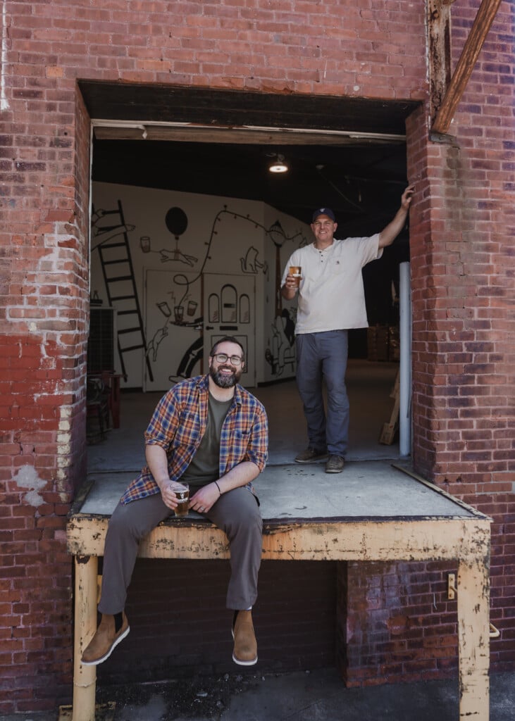 Two men, one sitting and one standing, smile and hold beers on the loading dock of a red brick mill building.