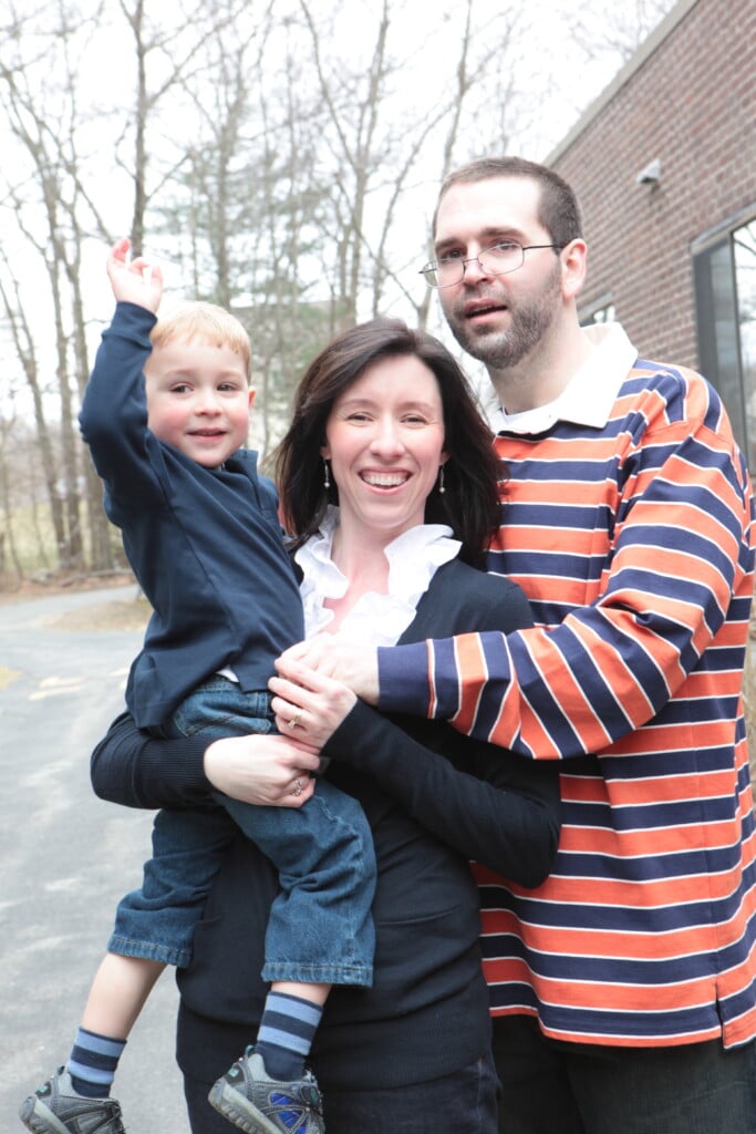 A woman holding a toddler and a man pose for a family photo outside.