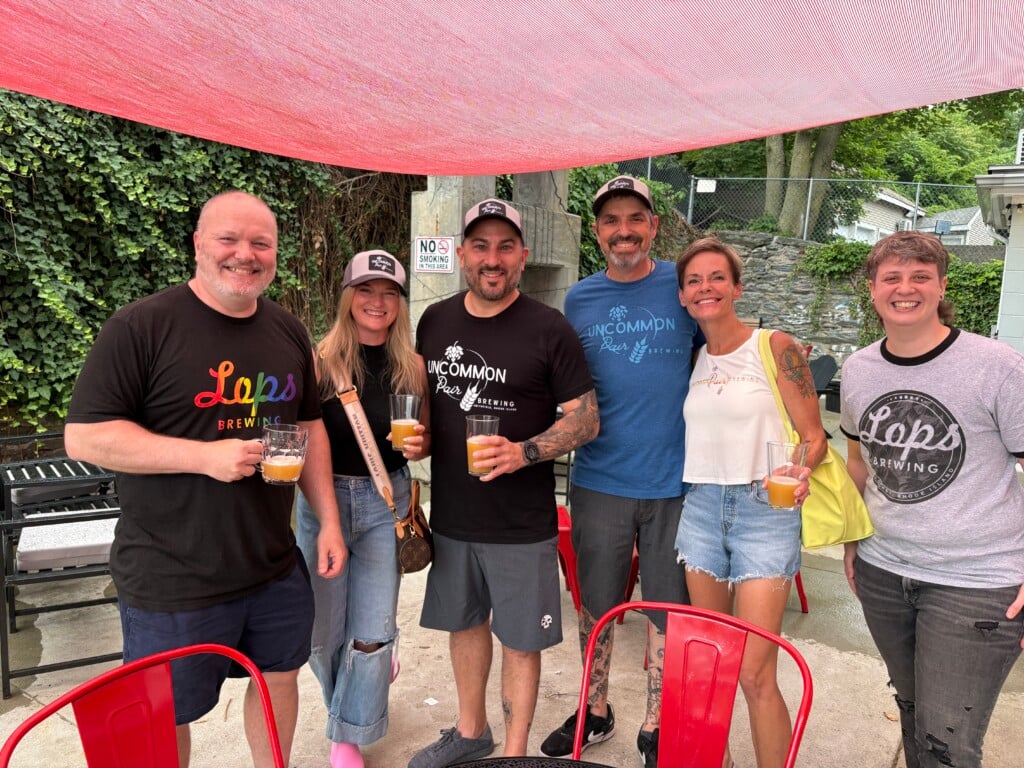 A group of people holding beers stand on an outdoor patio.